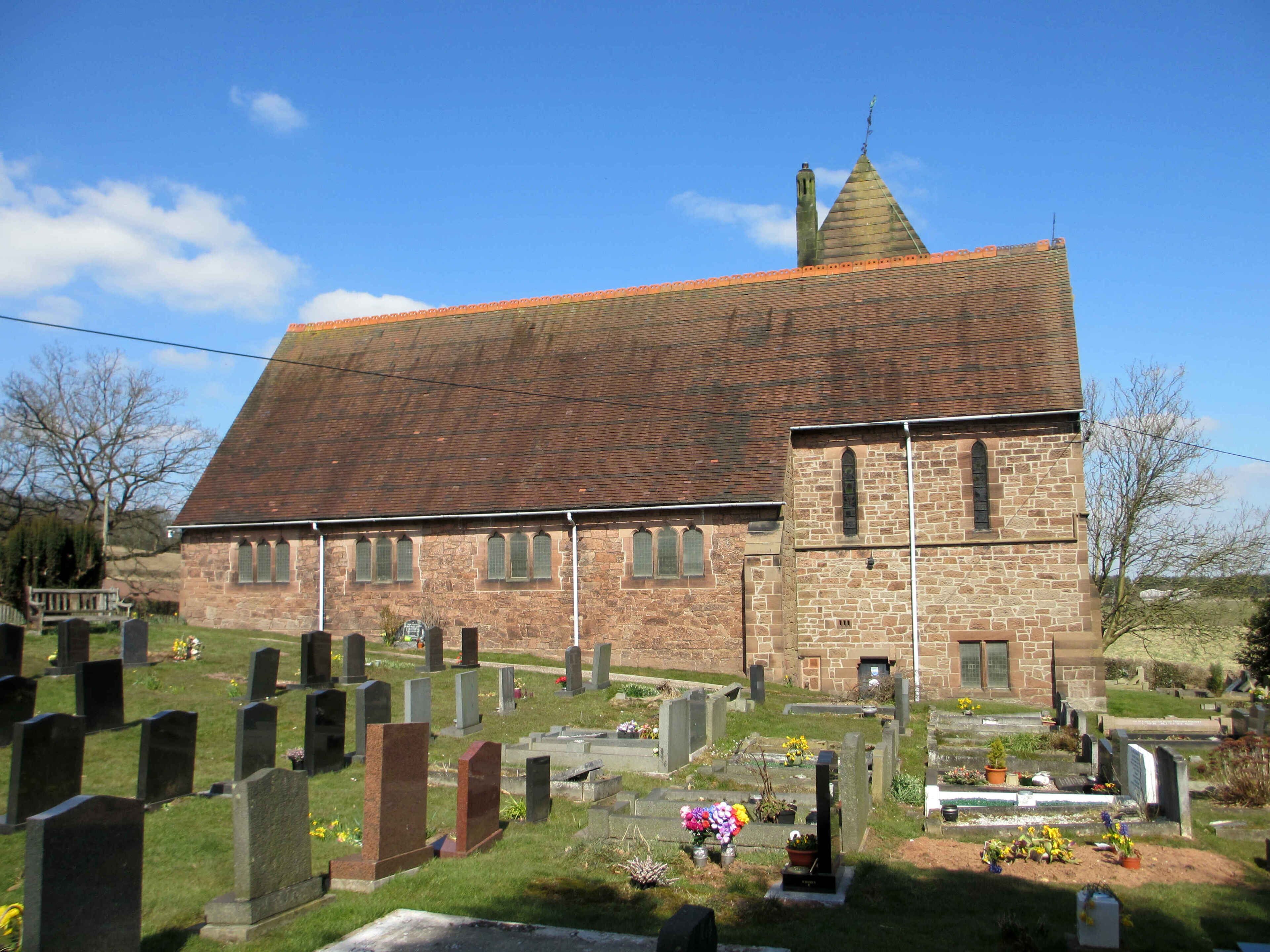A church with a red-tiled roof and stone walls, surrounded by a cemetery with gravestones and flowers.