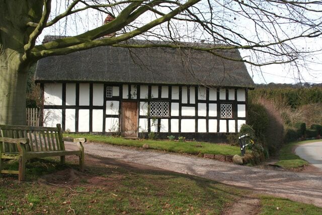 A traditional half-timbered house with a thatched roof, a bench, and a pathway leading to it.