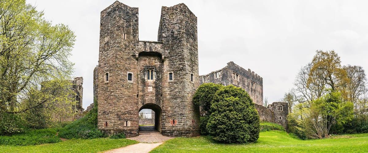 A historic stone castle with two prominent towers, surrounded by lush greenery and a well-maintained lawn.