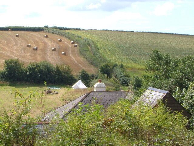 Roofs at Chilley. Tent, farmhouse, outhouse at Lower Chilley Farm from the bridleway which ends here. The contining green lane southwestwards is in the background, climbing past the bales and maize.