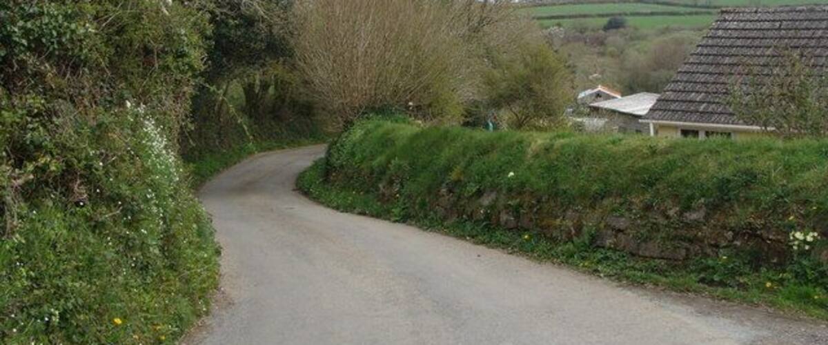 A narrow rural road curving through a countryside with greenery and a traditional house.