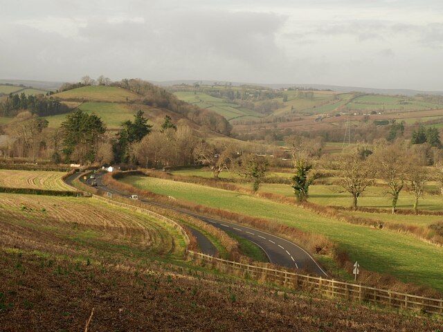 A384 below Huxham's Cross. The main road to the west of 1080828, as it sweeps round bends while descending to cross the River Dart. The prominent hill on the left is 156150.