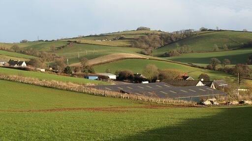 Longcombe. Taken from the edge of the car sales establishment shown in 1143712. The hamlet is on the northern slopes of one of the valleys that runs down to 914221. The slopes beyond, with Broomball Plantation occupying a cleft, are in SX8459, and Windmill Hill Clump, on the central hilltop, is in SX8559.