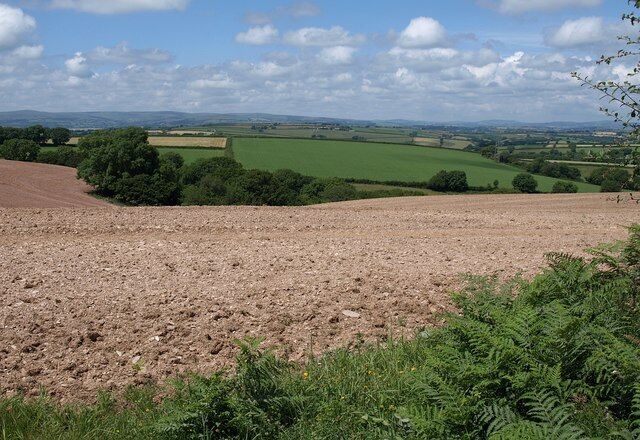 A rural landscape with fields, trees, and rolling hills under a partly cloudy sky.