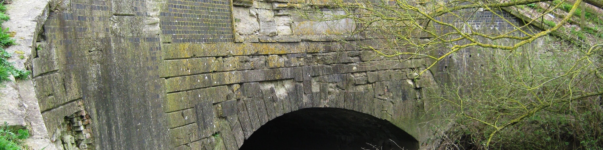 Uma ponte de pedra com um túnel embaixo, cercada por vegetação e uma cerca de madeira.