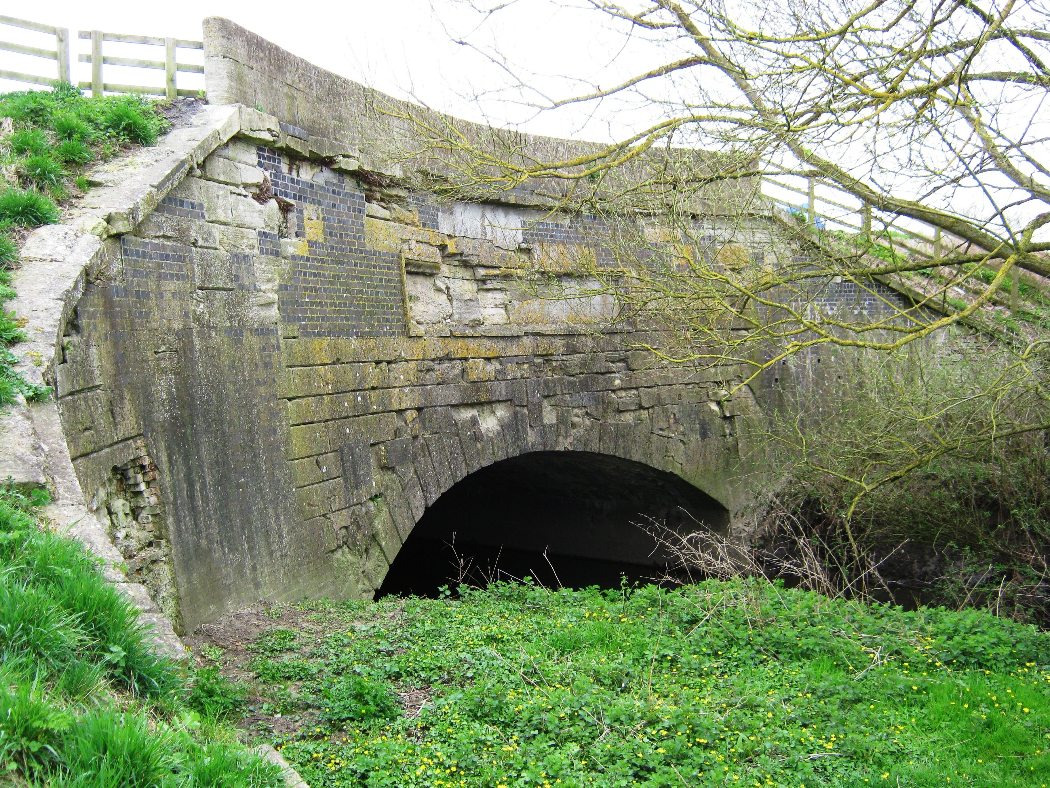 A stone bridge with a tunnel underneath, surrounded by greenery and a wooden fence.