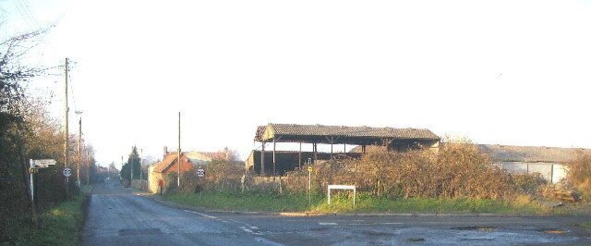 A rural road with a large building on the right and a smaller structure in the distance.