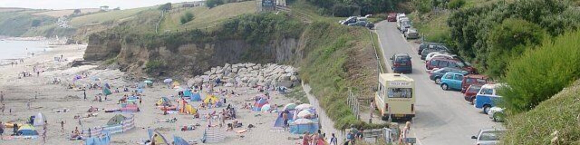 Uma praia com tendas e pessoas, uma estrada com carros e casas ao fundo.