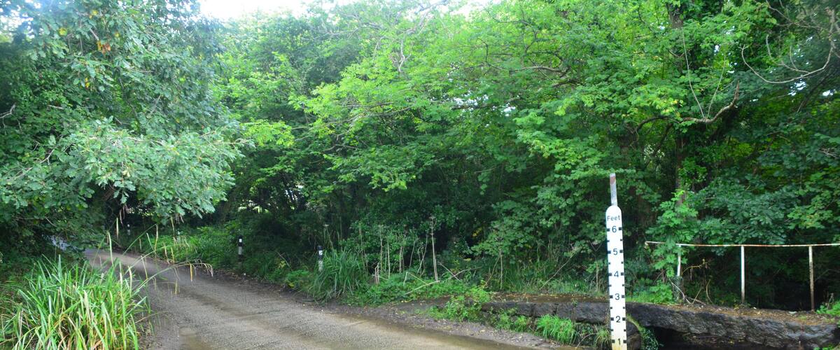 A narrow road crossing a shallow stream with a height marker.