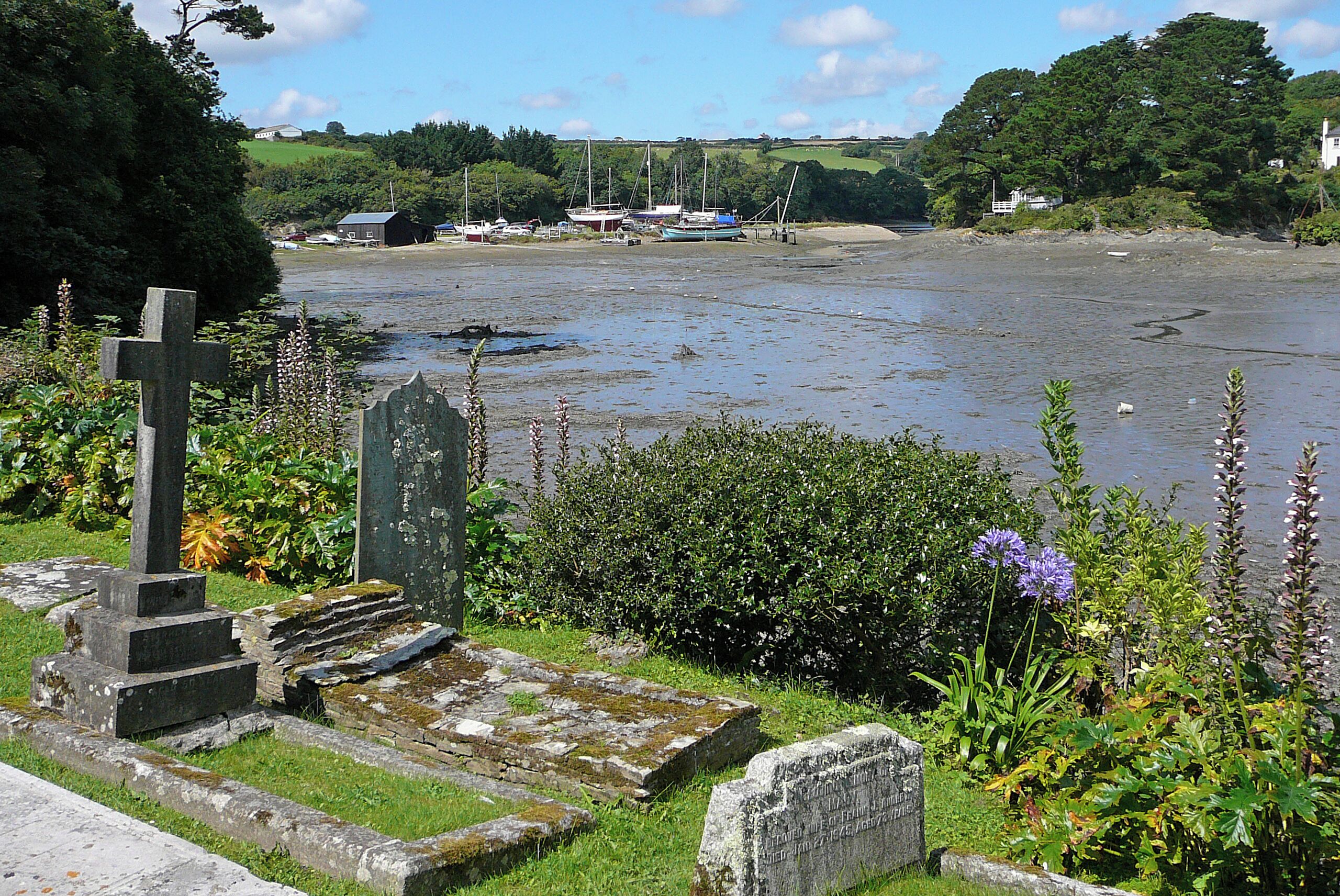 A cemetery with a prominent cross and gravestones, a river with boats, and a backdrop of greenery and houses.