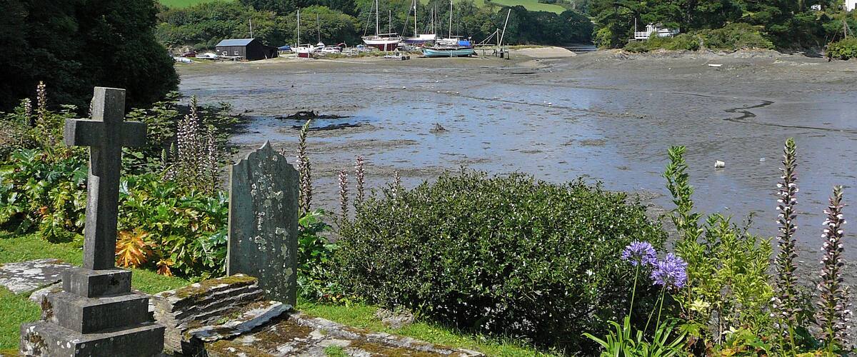 A cemetery with a prominent cross and gravestones, a river with boats, and a backdrop of greenery and houses.