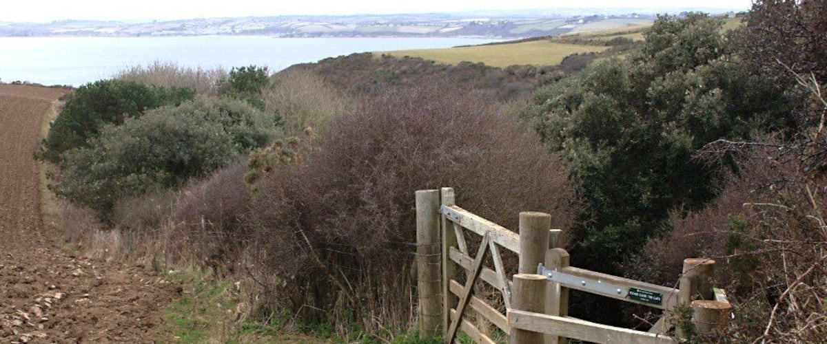A wooden gate at the entrance of a rural path with a view of a lake and hills in the background.