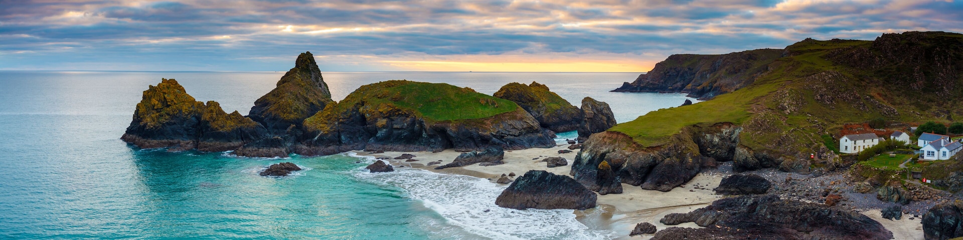 Paisagem costeira com rochas, uma praia de areia e casas ao fundo.