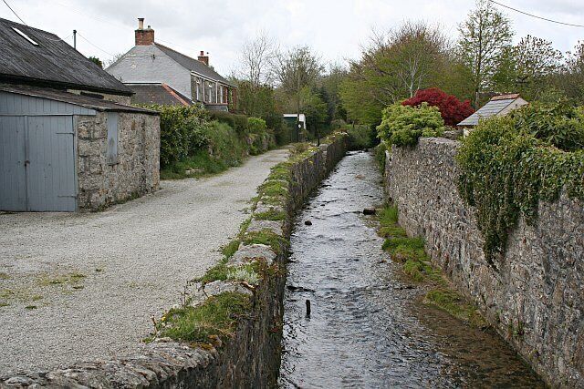 A narrow stone-lined water channel in a rural setting with houses and greenery.
