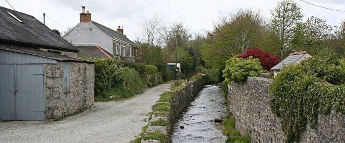 A narrow stone-lined water channel in a rural setting with houses and greenery.