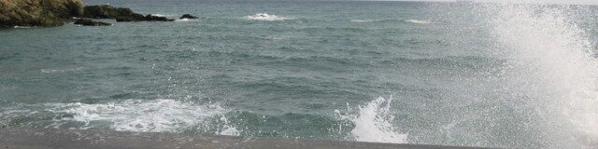 Um muro de concreto com ondas batendo e um penhasco rochoso ao fundo.