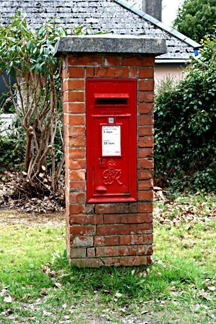 A red post box with a sign, surrounded by grass and trees.