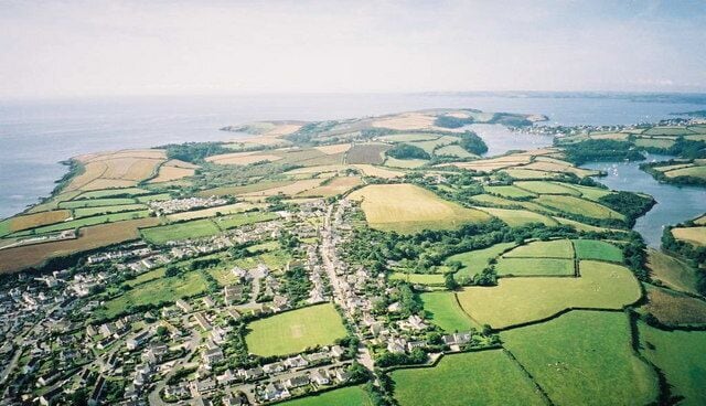 An aerial view of a coastal town with green fields, a river, and a sea in the background.