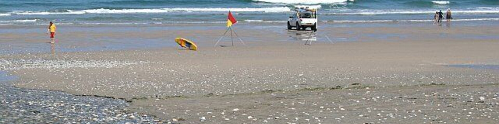 Una playa pedregosa con una torre de salvavidas, una bandera de salvavidas y un camión blanco.
