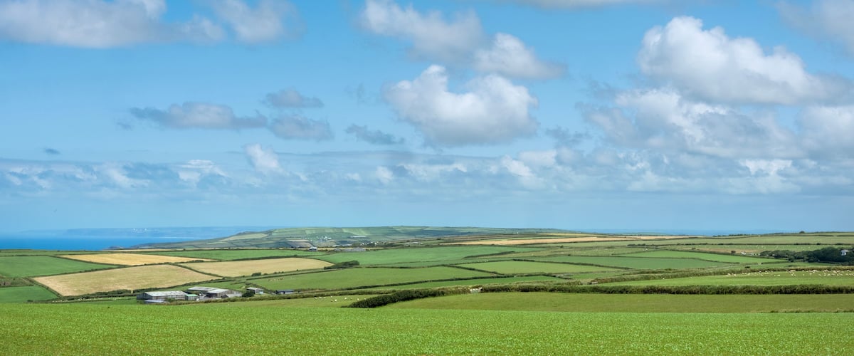 A rural landscape with green fields, scattered buildings, and a distant view of the sea under a partly cloudy sky.