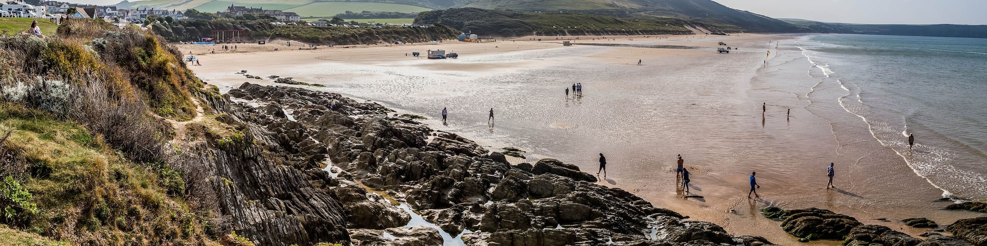 A coastal landscape with a sandy beach, rocky shoreline, and rolling hills in the background.