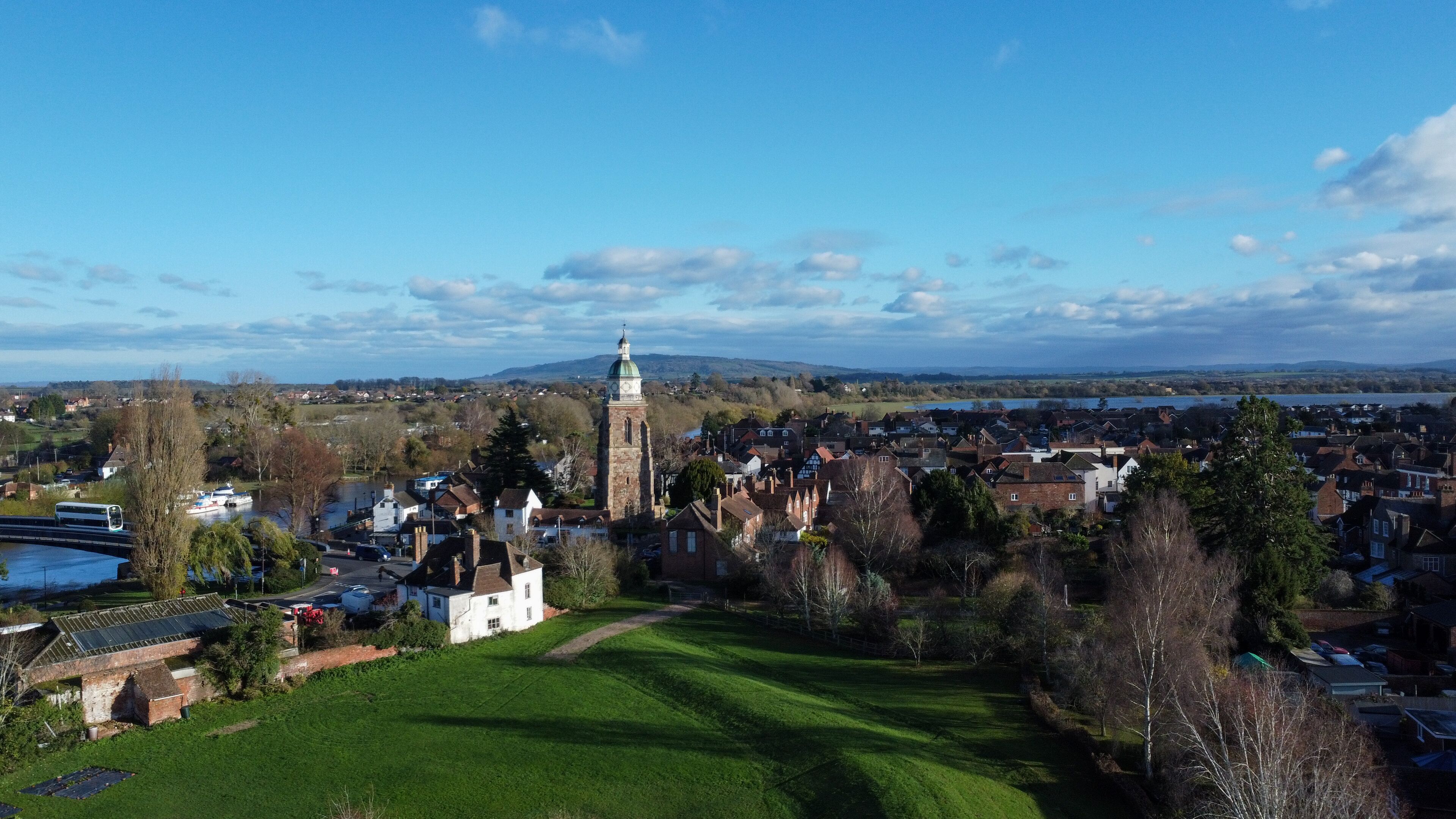 A town with a prominent clock tower, a river, and surrounding greenery.