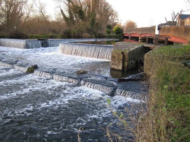 River Colne: Overspill weir from the Grand Union Canal. The Grand Union Canal is beyond the towpath footbridge on the red girder on the right. Water flows out of the canal, under the footbridge, and over the weir into the River Colne.