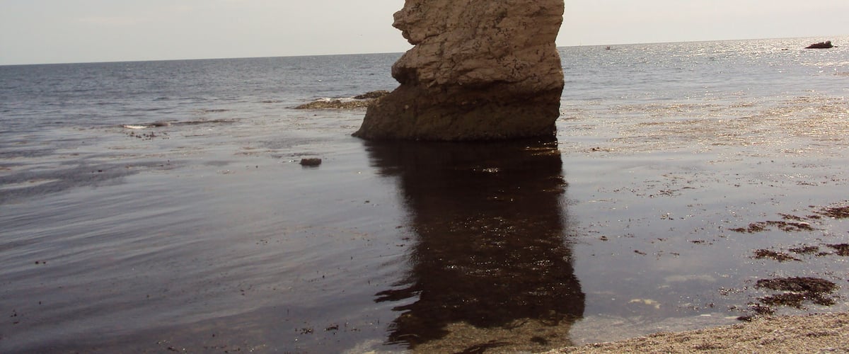 A large rock formation on a beach with calm water and a clear sky.