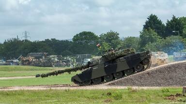 A military tank on a dirt track with a grassy field and trees in the background.