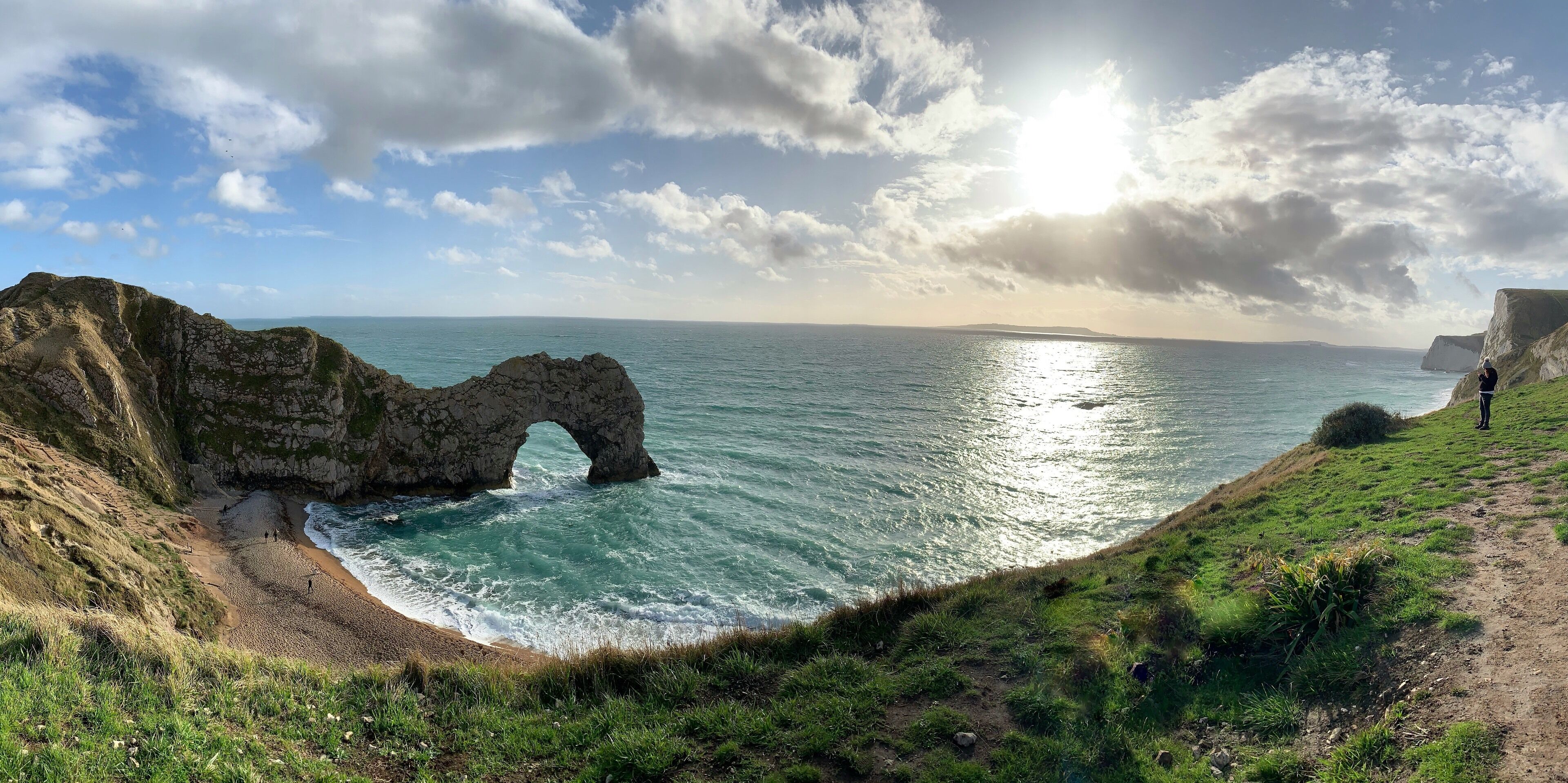 A coastal landscape with a prominent rock arch formation, a grassy path, and the ocean.