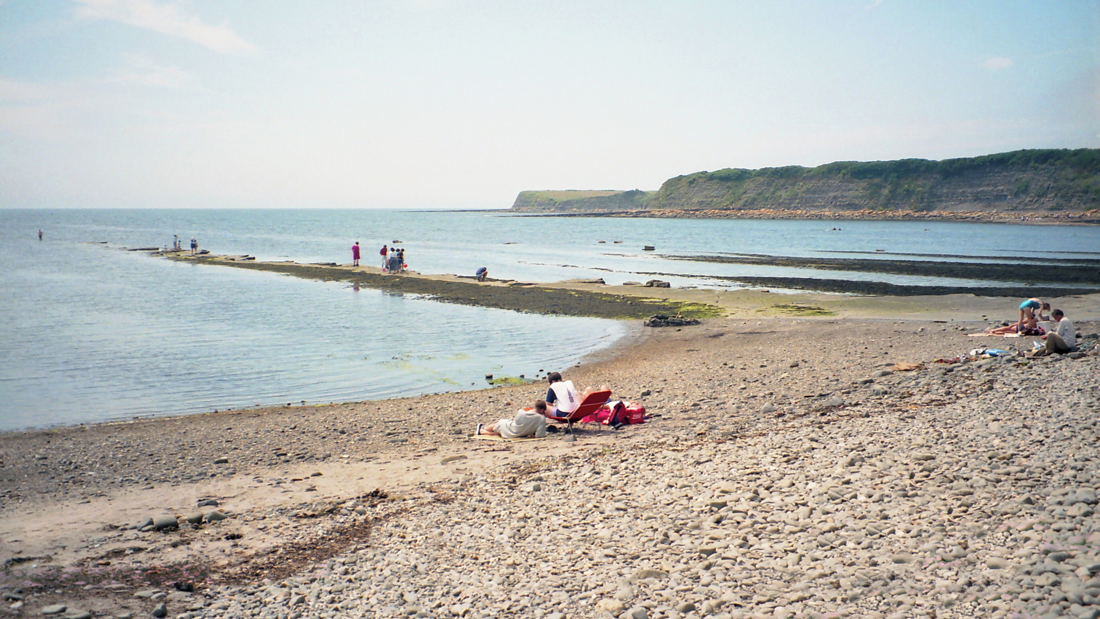 A pebbly beach with people relaxing and a distant cliff.