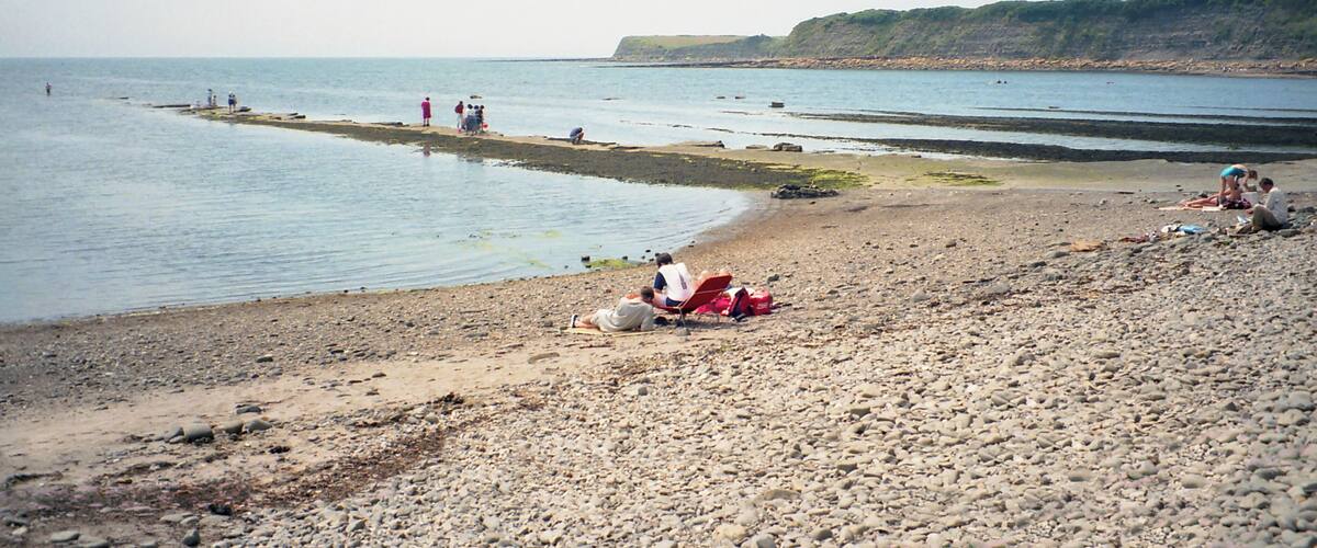A pebbly beach with people relaxing and a distant cliff.