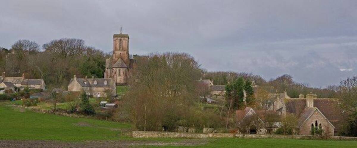 A historic church with a tall tower, surrounded by traditional stone houses and a green field.