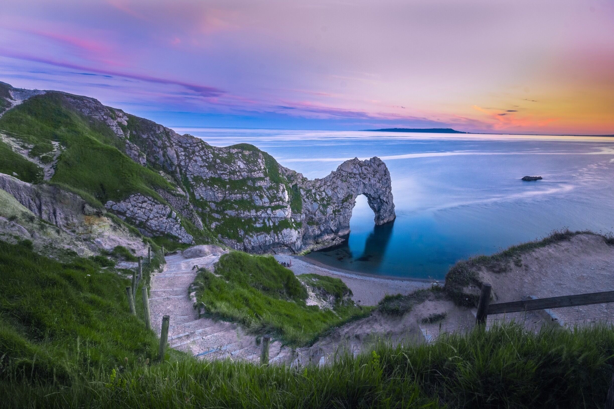 A coastal landscape with a prominent rock arch formation, a clear sky, and a calm sea.