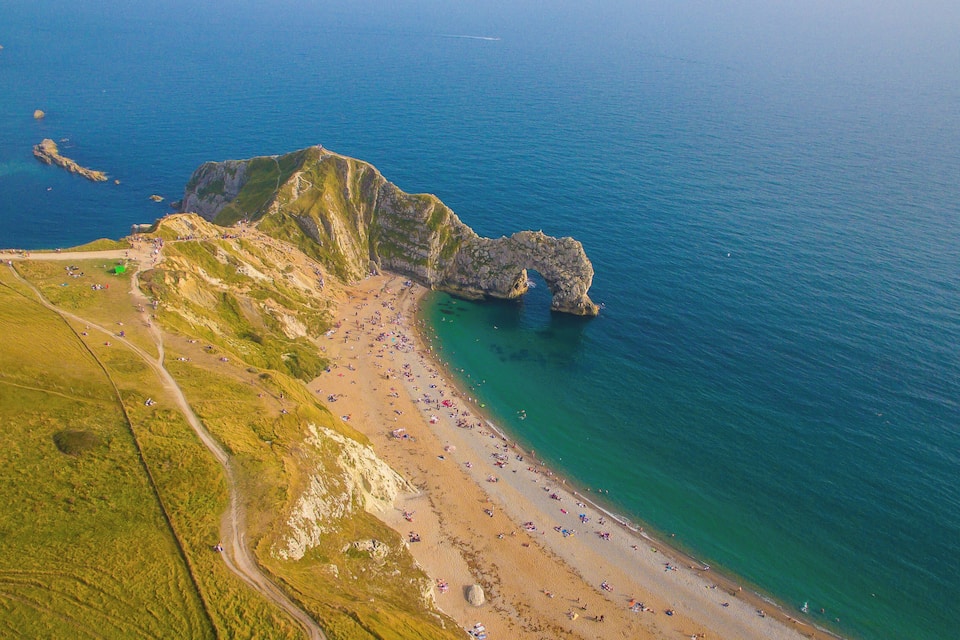 An aerial view of a coastal area with a rocky cliff, a sandy beach, and a clear blue sea.