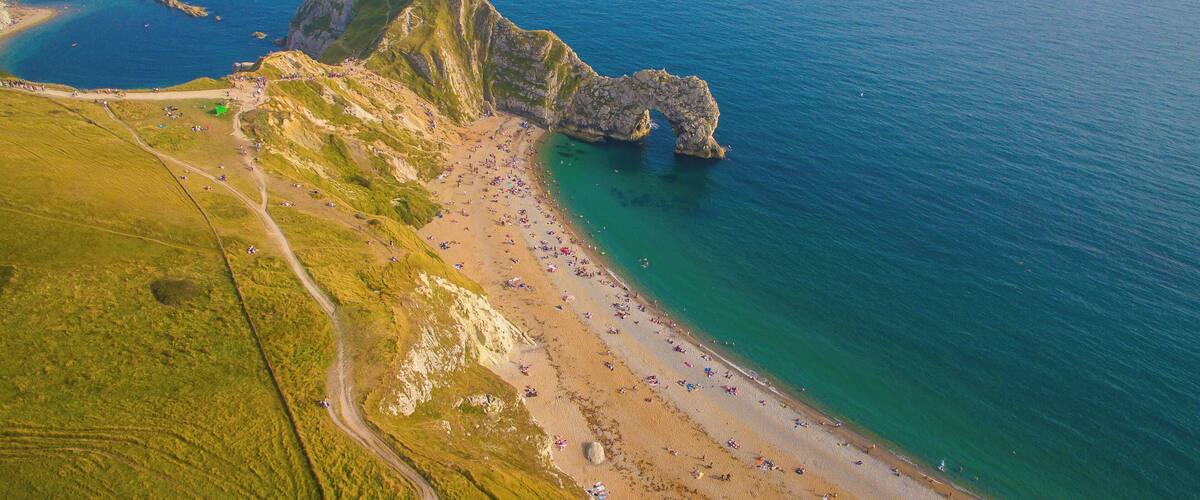 An aerial view of a coastal area with a rocky cliff, a sandy beach, and a clear blue sea.