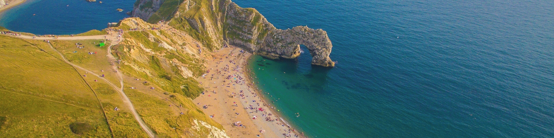 An aerial view of a coastal area with a rocky cliff, a sandy beach, and a clear blue sea.