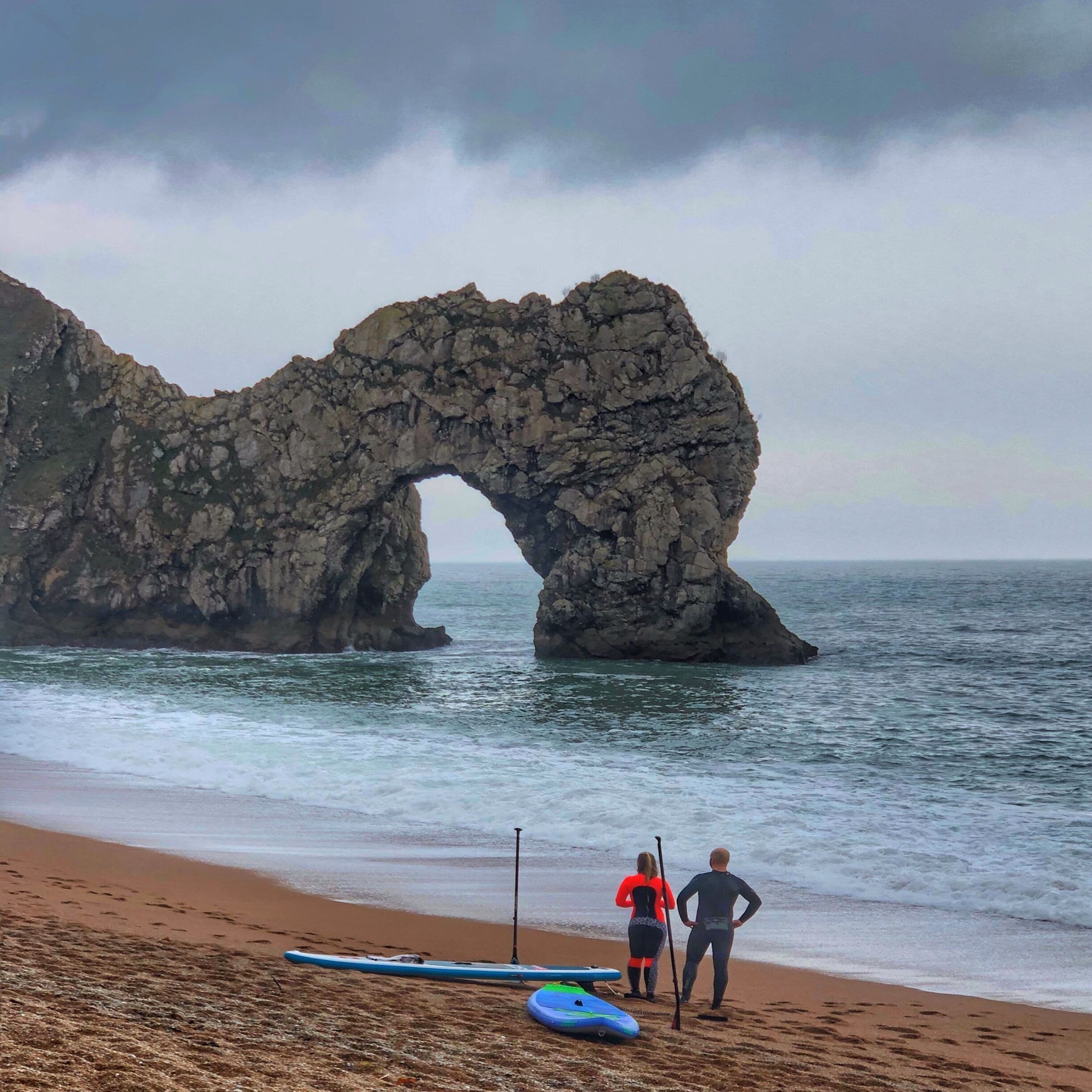 A rocky arch formation on a beach with two people standing nearby.