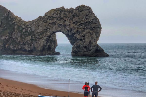 A rocky arch formation on a beach with two people standing nearby.