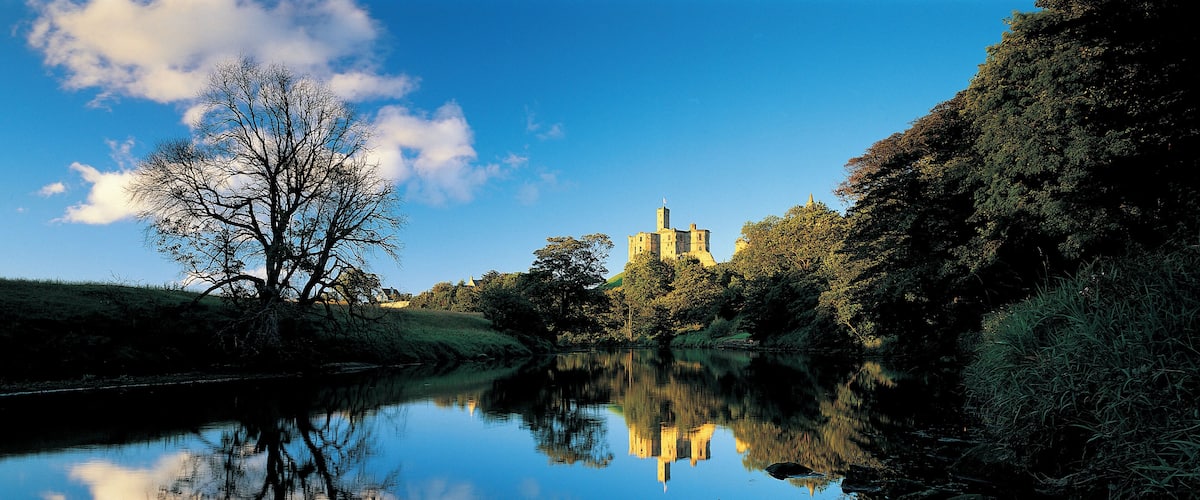 A castle reflected in a calm river, surrounded by trees and greenery.