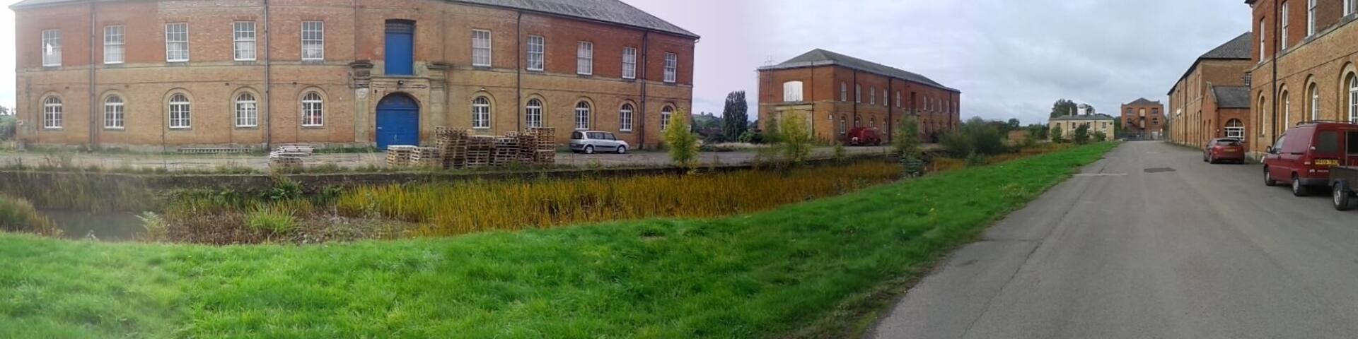 Former Ordnance Depot at Weedon Bec. The extensive barracks were begun in 1803 during the Napoleonic wars, but most of the buildings (including 12 powder magazines) have been demolished. This is a 180-degree panoramic shot and therefore the straight road appears as two roads. In fact it is parallel to the grass verge and canal in the foreground.