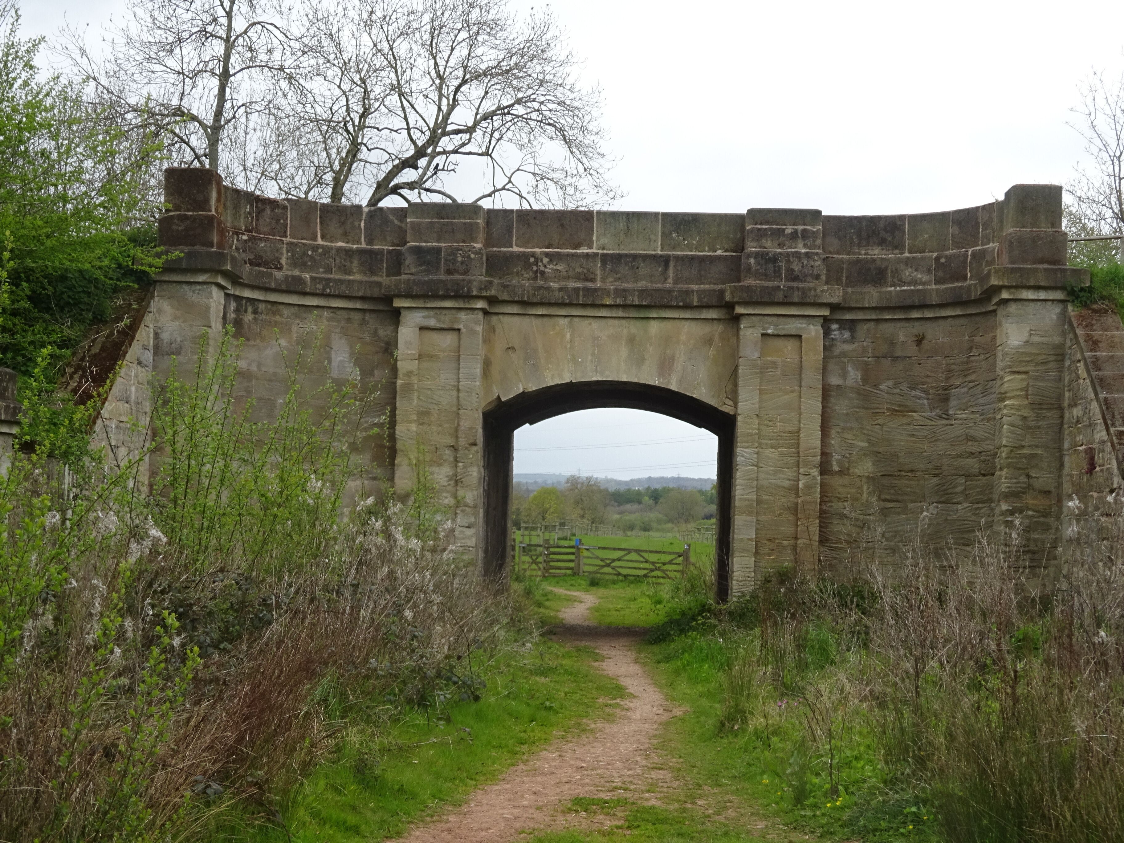A stone arch bridge in a rural setting with a dirt path leading through it.