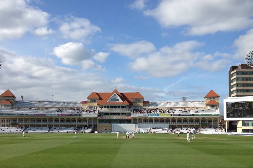 A cricket stadium with a large field, tiered seating, and a scoreboard.