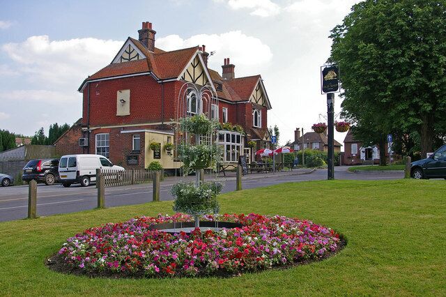 A red brick building with a Tudor-style roof, a flower bed, and a street with cars and a white van.