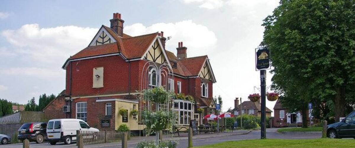 A red brick building with a Tudor-style roof, a flower bed, and a street with cars and a white van.