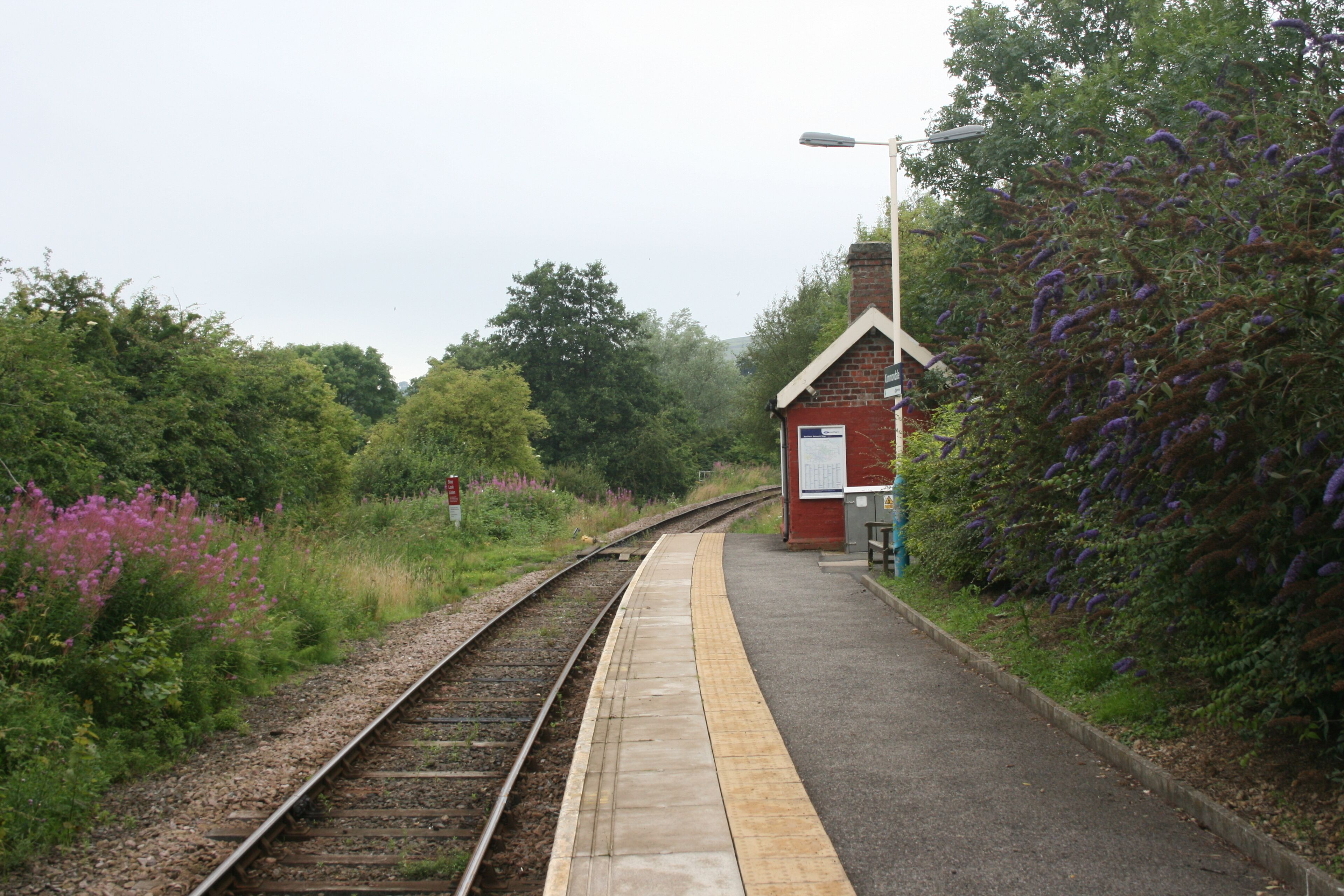 A railway platform with a red building, surrounded by greenery and purple flowers.