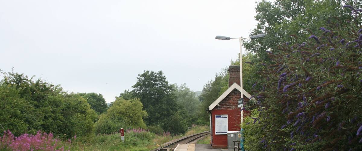 A railway platform with a red building, surrounded by greenery and purple flowers.