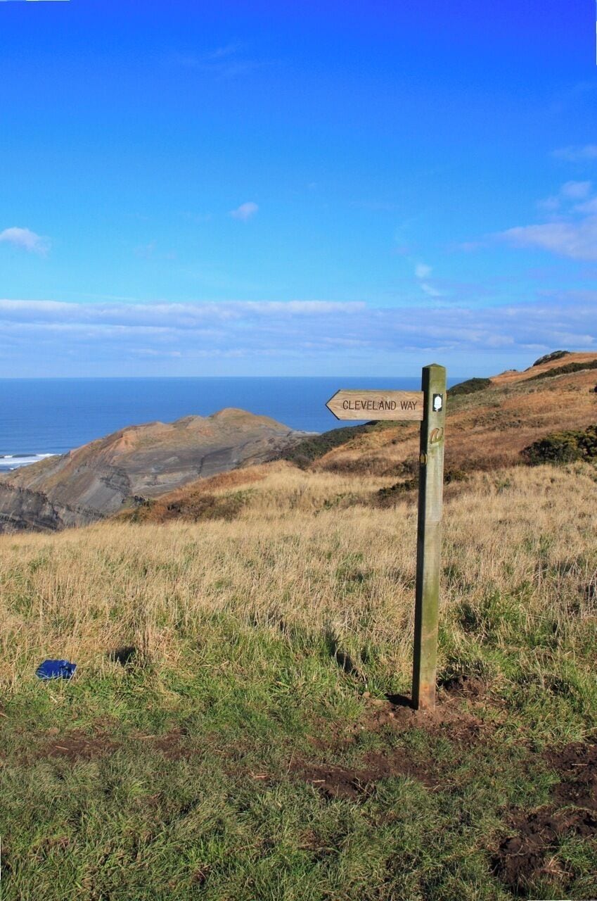 A wooden signpost with 'Cleveland Way' pointing to the right, overlooking a coastal landscape with grassy hills and the ocean in the background.