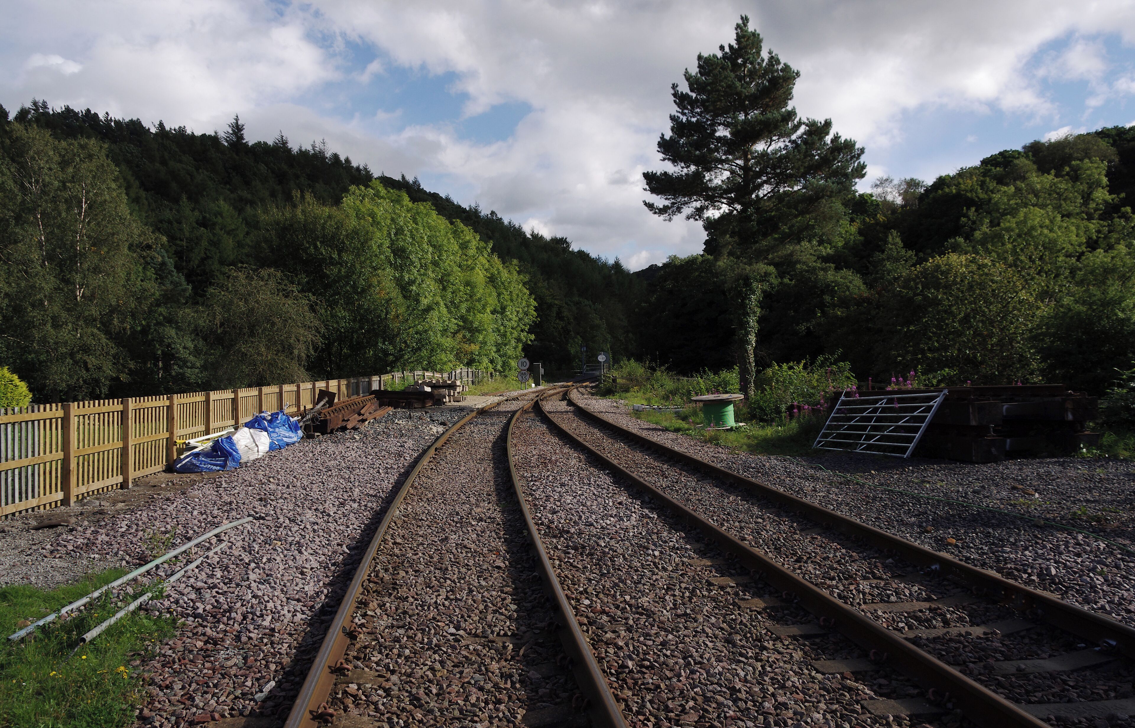 Railway tracks curving through a forested area with a wooden fence and some construction materials on the side.