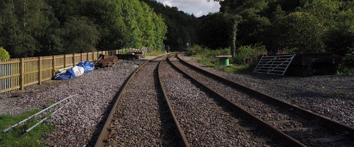 Railway tracks curving through a forested area with a wooden fence and some construction materials on the side.